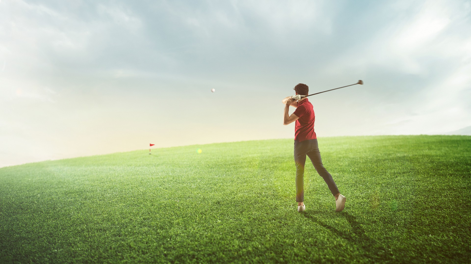 Young man, golf player in red shirt taking a swing, playing golf on field at sunny summer day, outdoors.